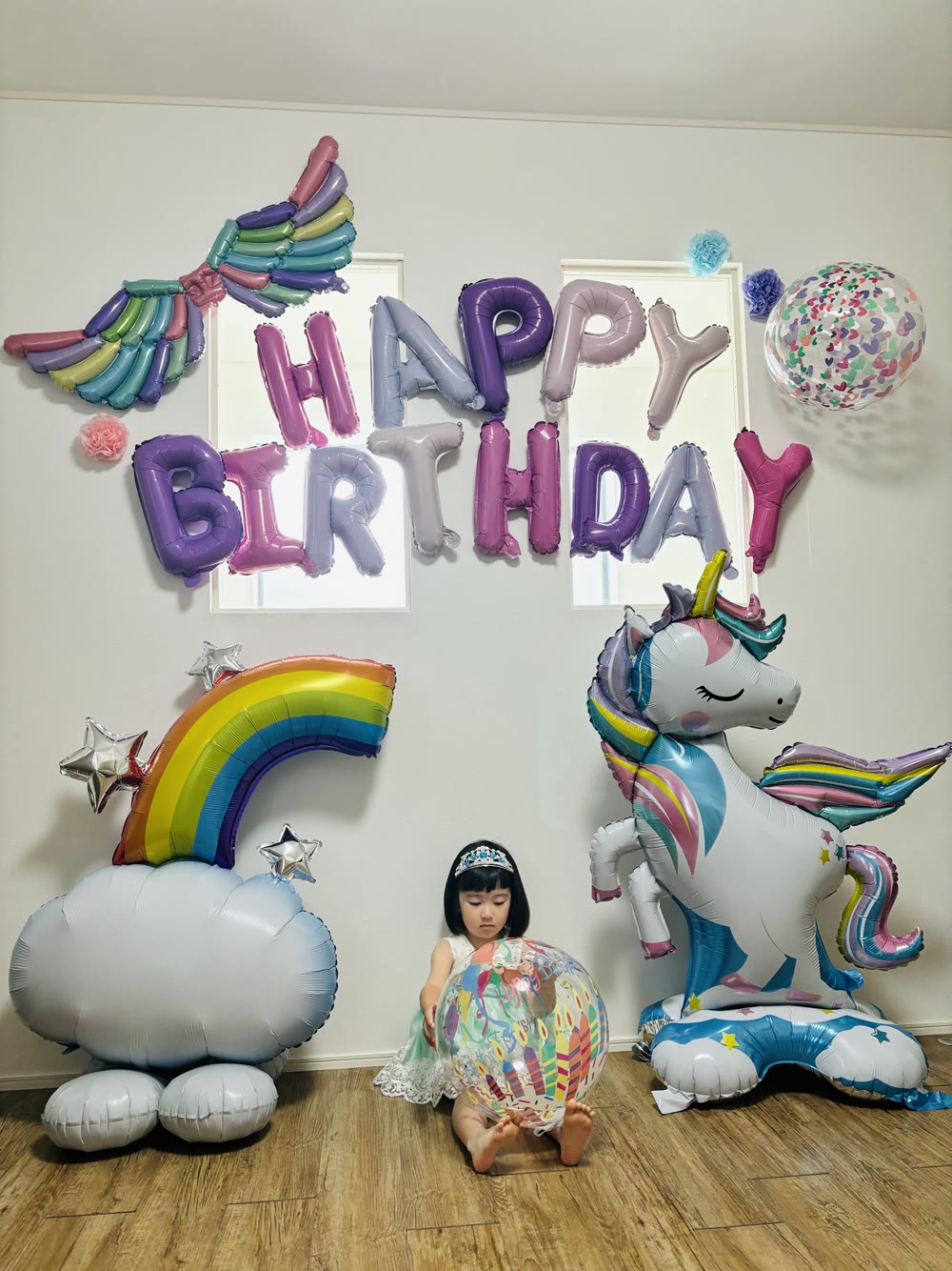 自立式の大きなユニコーンと虹のバルーンのあいだに座るティアラの女の子