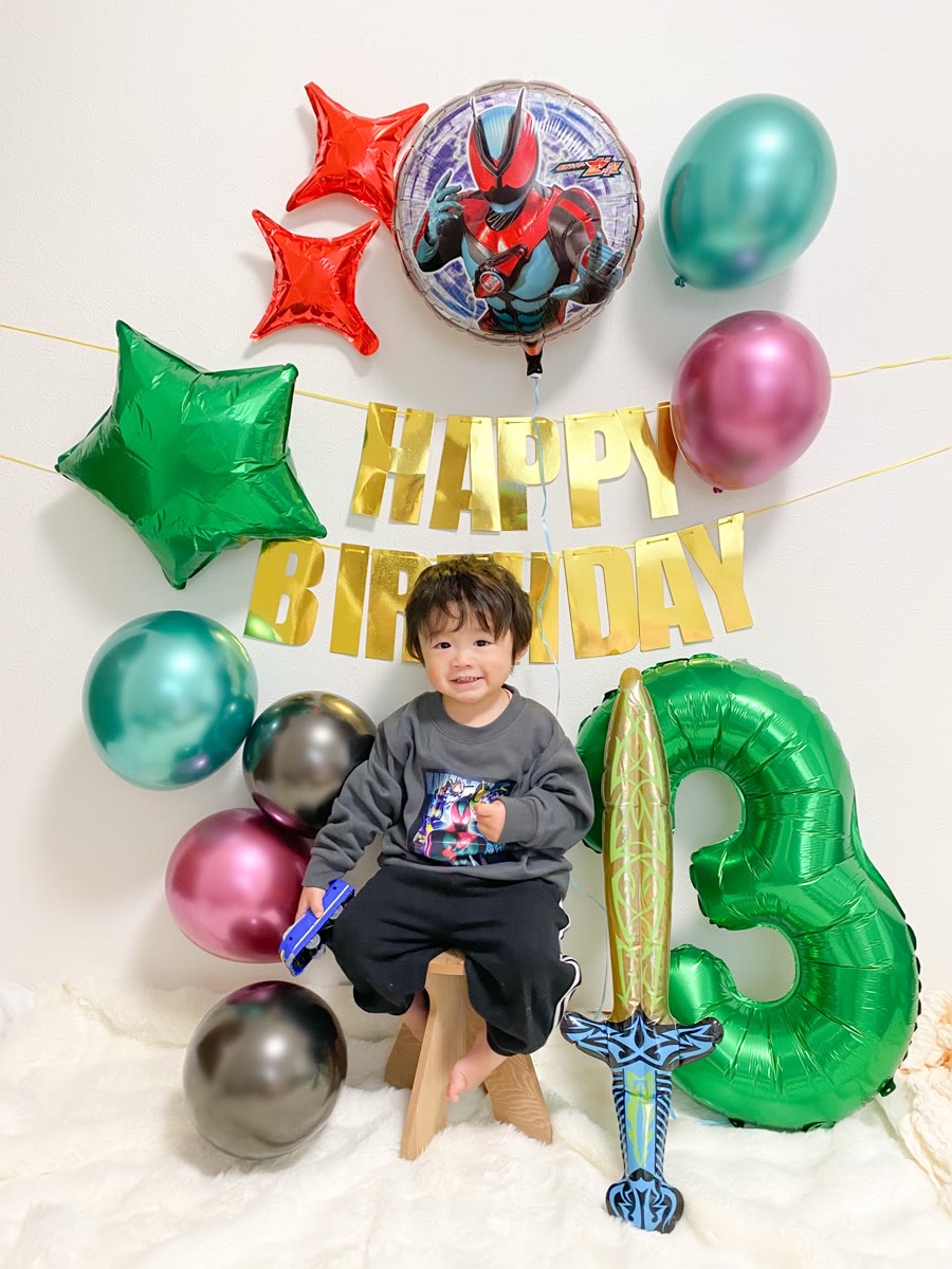 仮面ライダーゼッツの丸形バルーンと剣のバルーン、緑の数字3バルーン、金のHAPPY BIRTHDAYガーランド、メタリックのゴム風船の前でスツールに座って笑う男の子