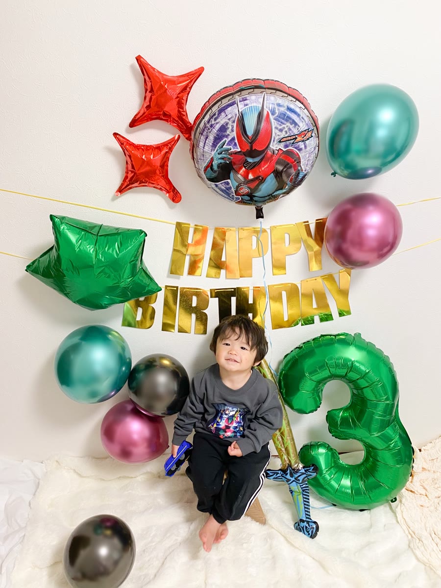 仮面ライダーゼッツのバルーンと数字3、剣のバルーンのそばでミニカーを持って座る男の子。メタリックの赤・青・黒のゴム風船